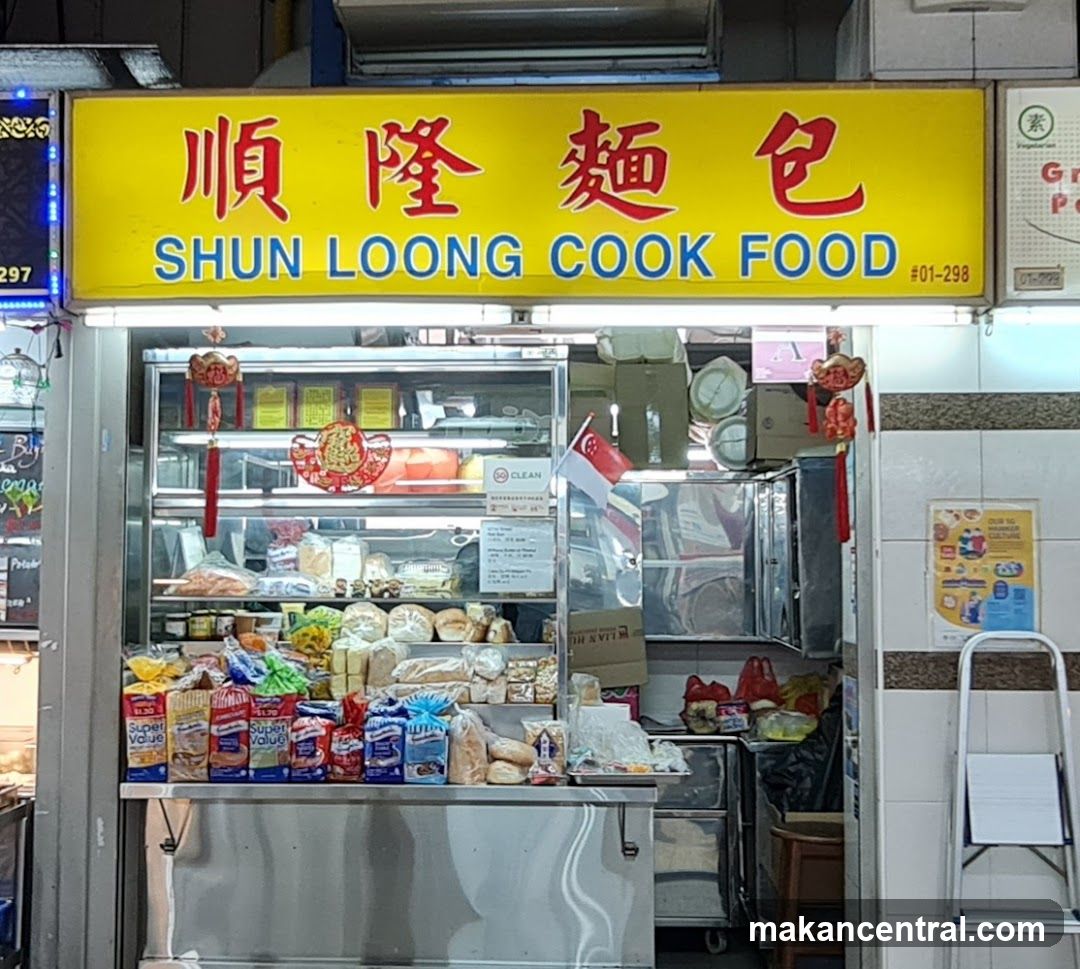 A storefront with a bright yellow sign featuring red Chinese characters "順隆麵包" and blue English text "SHUN LOONG COOK FOOD #01-298". The shop displays baked goods in refrigerated glass cases, packaged products on the counter, and Singaporean flags. Red decorative elements hang from the sign. A ladder is visible to the right.
