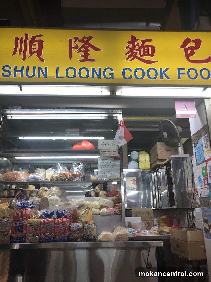 Yellow sign with red Chinese characters "順隆麵包" and blue English text "SHUN LOONG COOK FOOD" above a display counter filled with packaged breads and baked goods. A small Singapore flag is visible inside the shop. The interior includes a fan, stacked boxes, and a stainless steel display case.