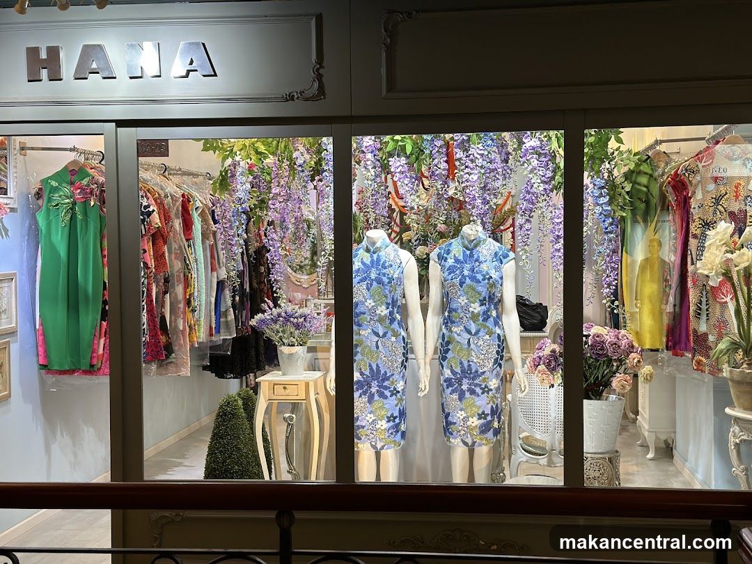 Storefront with 'HANA' sign above glass display windows showing mannequins in floral qipao dresses, racks of colorful garments, hanging purple wisteria flowers, and decorative plants. Interior lighting illuminates the display. A wooden railing is visible in foreground.