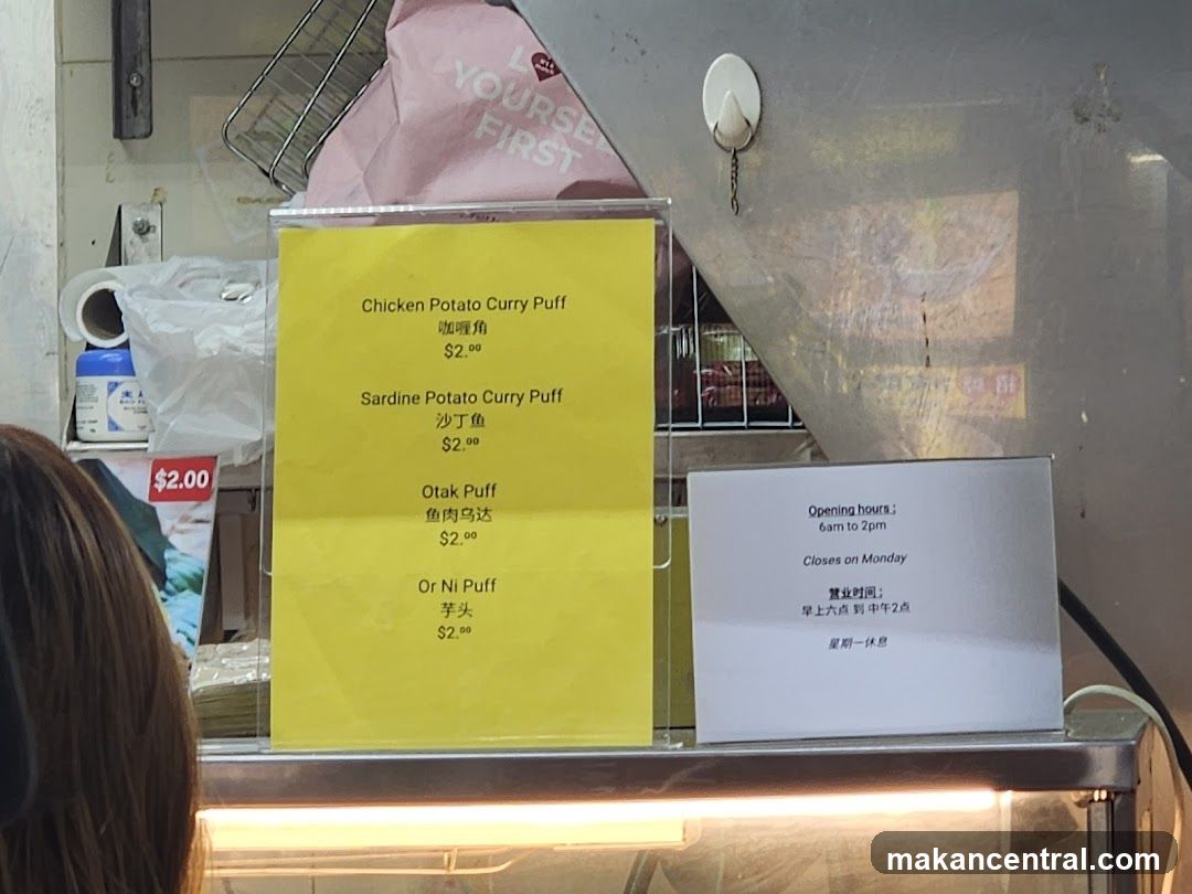 Yellow menu board displaying four curry puff options with prices in USD. Adjacent white sign shows opening hours and closing on Monday.