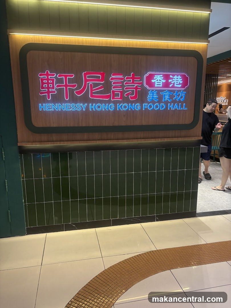 A storefront sign with illuminated Chinese characters '軒尼詩香港美食坊' and English text 'HENNESSY HONG KONG FOOD HALL'. The sign is mounted on a wooden panel above a green tiled base, under warm lighting. A person walks by to the right.