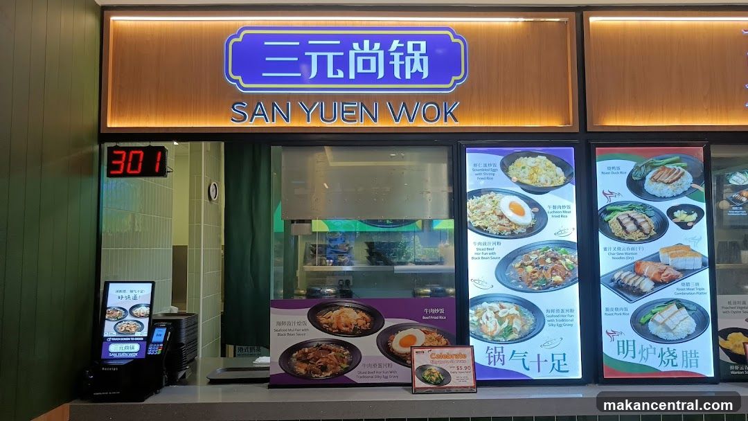 Exterior view of a restaurant entrance with illuminated sign reading '三元尚锅' and 'SAN YUEN WOK'. A digital clock displays '301', likely indicating time or order number. Behind the counter, a glass window shows kitchen area. To the right are two vertical menu boards featuring images of dishes like fried rice, seafood bowls, and roasted meats. A small promotional card is placed on the counter.