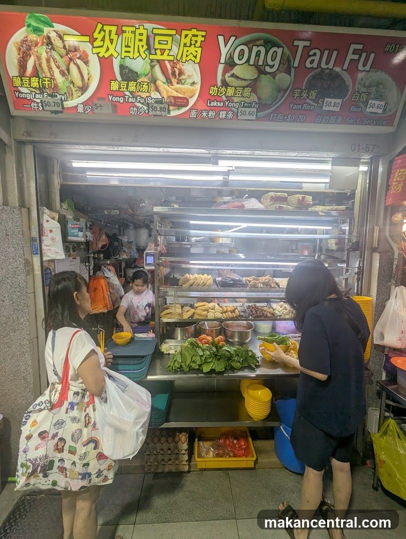 Exterior view of a food stall with bright red signage displaying '一级酿豆腐 Yong Tau Fu'. Customers are at the counter, one holding a bowl, another carrying bags. Inside, shelves hold prepared foods and fresh vegetables. A child is visible behind the counter.