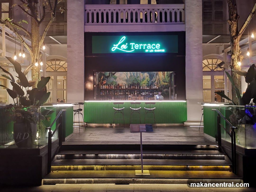 Nighttime exterior view of a bar with neon sign 'Lei Terrace by Lai Garden' above a green-lit bar counter. The entrance features steps leading up to the bar area, flanked by trees and glass planters. A bartender is visible behind the bar, which displays bottles against a jungle-themed mural. Ambient lighting highlights the structure's colonial-style architecture with arched windows and white pillars.