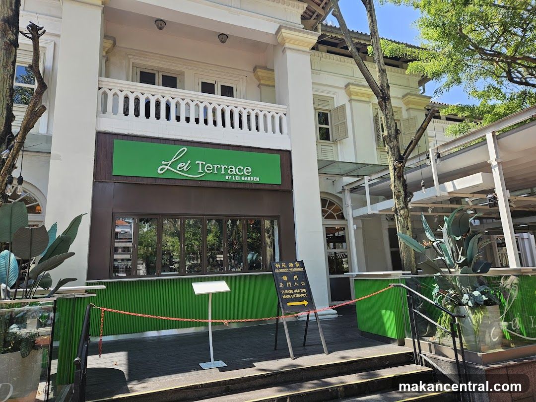 Exterior facade of a two-story building with white pillars and balcony, featuring a green sign with "Lei Terrace BY LEI GARDEN" above large windows. Green paneling lines the ground floor. A red rope barrier and directional sign indicate main entrance. Trees and potted plants flank the entrance.