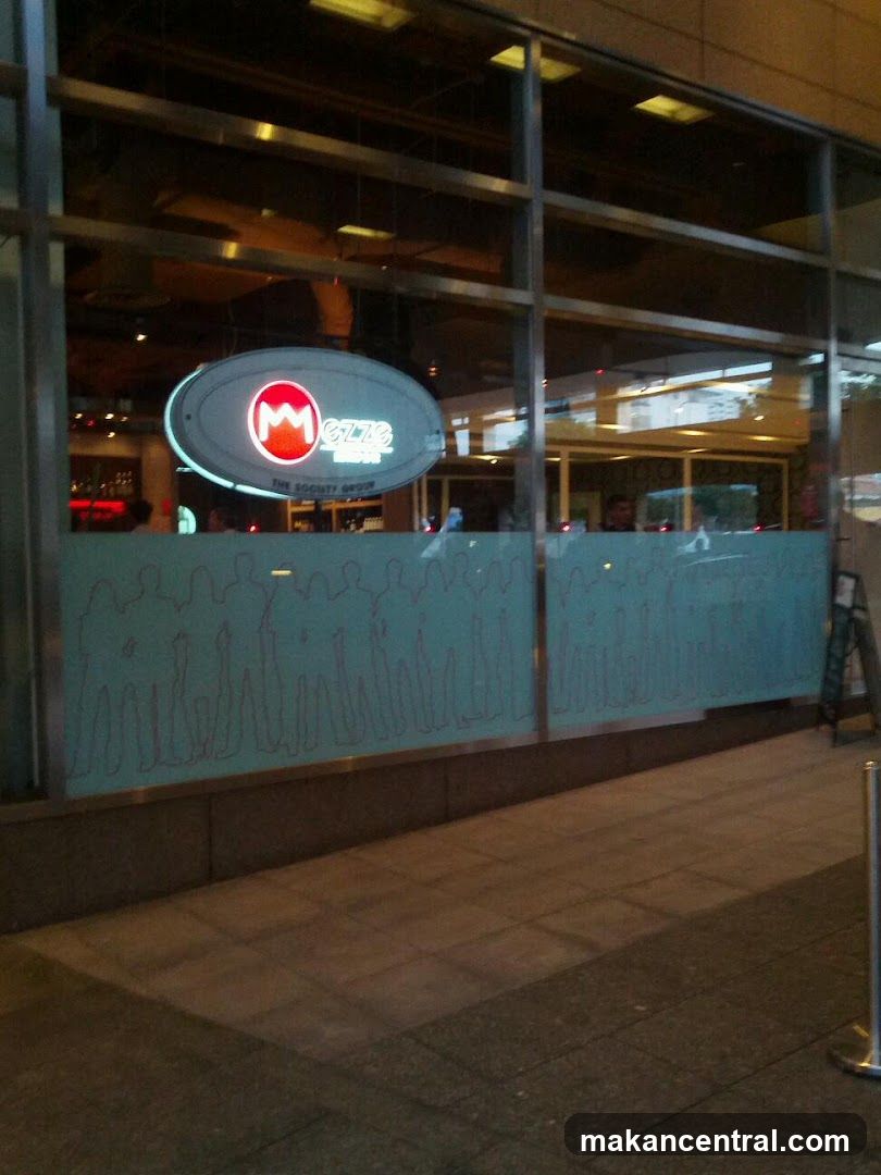 Exterior view of a modern storefront with large glass windows and a prominent oval-shaped illuminated sign featuring the logo 'M' and '6778'. The lower part of the window has a frosted blue panel with abstract line art. A metal stanchion is visible on the sidewalk to the right.