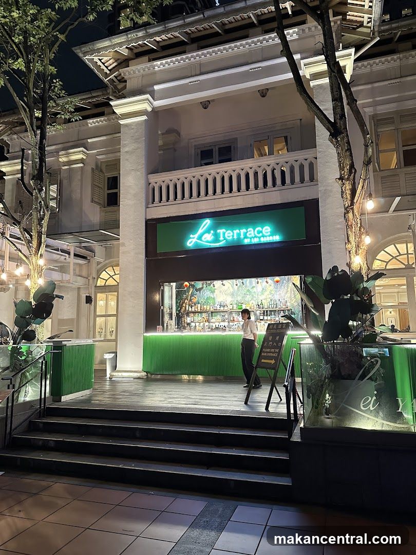 Nighttime exterior view of a two-story building with classical architecture, featuring a bright green neon sign reading 'Lei Terrace by the Gardens' above an open bar area. A staff member stands near the counter. Green accents and tropical plants flank the entrance. Stairs lead up to the shop front.