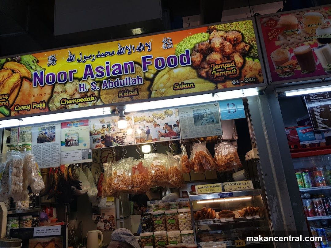 Storefront with large colorful signboard featuring 'Noor Asian Food' and owner name 'H.S. Abdullah'. Sign includes Arabic script and images of food like tempur temput and chempedak. Below the main sign, hanging bags of snacks and a display case with baked goods are visible. A refrigerator to the right holds canned drinks. The shop appears busy with interior lighting and promotional posters on walls.