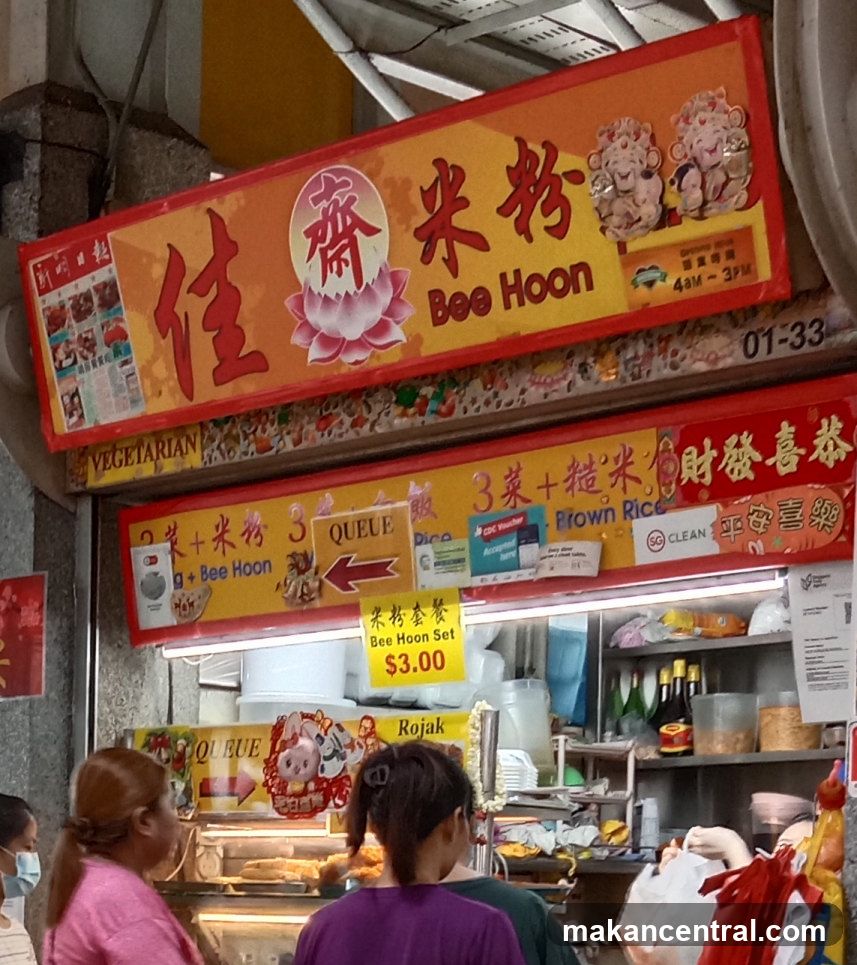 A storefront with a large red and yellow sign featuring Chinese characters '佳齋米粉' and English text 'Bee Hoon'. The sign includes decorative elements like lotus flowers and figurines. Below the main sign, there are smaller signs indicating vegetarian options, queue directions, and a clean certification logo. A small yellow price tag reads 'Bee Hoon Set $3.00'. Customers are visible in front of the stall.