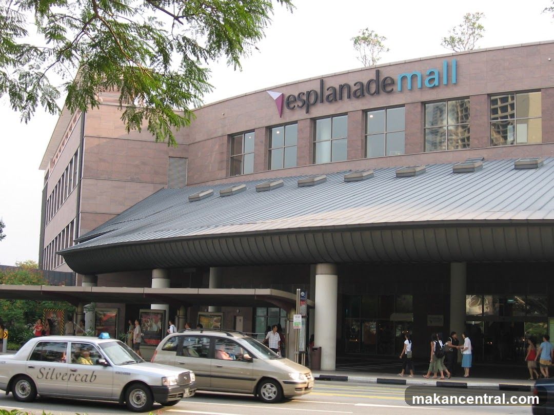 Exterior view of Esplanade Mall's main entrance with a modern facade featuring large windows and a curved metal roof overhang. The mall's name is prominently displayed in blue and black lettering on the upper part of the building, accompanied by a triangular logo. Several people are seen walking near the entrance, and two taxis are parked or moving along the street in front. Trees partially frame the top left corner of the image.