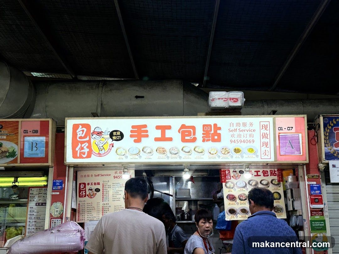 A storefront with a bright white sign featuring red Chinese characters '手工包點' and English text 'Self Service'. The sign includes images of dishes and contact number 94594069. Below the main sign, there are smaller menus and promotional materials on the walls. Customers are seen inside or near the counter. The ceiling is dark meshed metal, and adjacent stalls are partially visible.