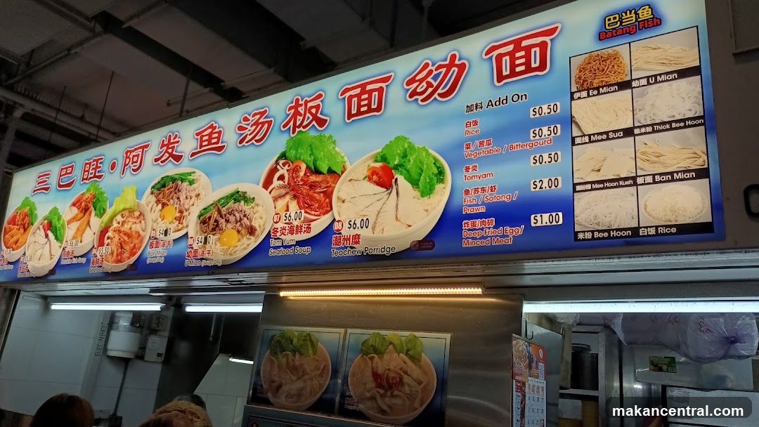 A brightly colored menu board displaying noodle dishes with prices in Singapore dollars. The board features images of bowls with ingredients like fish, vegetables, and egg. Add-on options include rice, vegetable, tofu, and deep-fried egg/minced meat. Also shows variations like batang fish noodles and thick bee hoon.