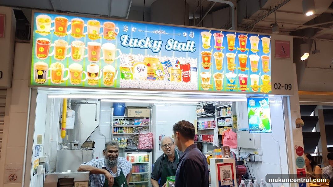 A brightly lit stall with a large illuminated sign displaying the name 'Lucky Stall' and images of various beverages. The storefront is open, revealing interior shelves stocked with drinks and snacks. Two men are conversing near the counter, while another staff member stands behind it. A QR code payment station is mounted on the right side.