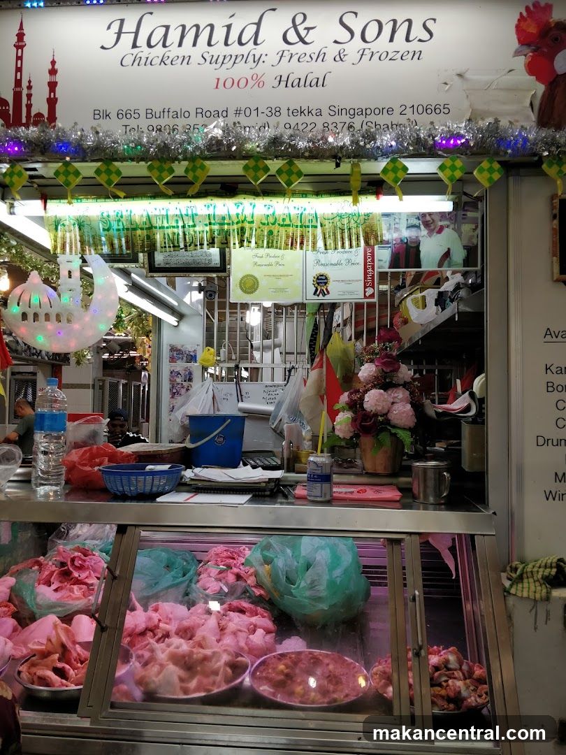 Storefront with sign reading 'Hamid & Sons Chicken Supply: Fresh & Frozen 100% Halal' at Blk 665 Buffalo Road #01-38 tekkas Singapore 210665. Decorated with festive green and yellow checkered flags, a crescent moon light fixture, and a rooster emblem. Display case shows raw chicken in plastic bags under glass. Behind counter are water bottle, blue bucket, flower arrangement, and framed photo of smiling people. Signage includes 'Selamat Hari Raya', halal certification, and Singapore license plaque.