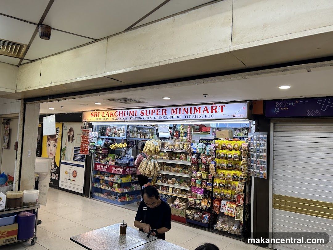 Storefront with bright red signage reading 'SRI LAKCHUMI SUPER MINIMART' above the entrance. Shelves stocked with snacks, drinks, and other goods are visible inside. A customer sits at a table outside the shop. The ceiling shows signs of wear. Adjacent to the store is a shuttered unit with purple branding.