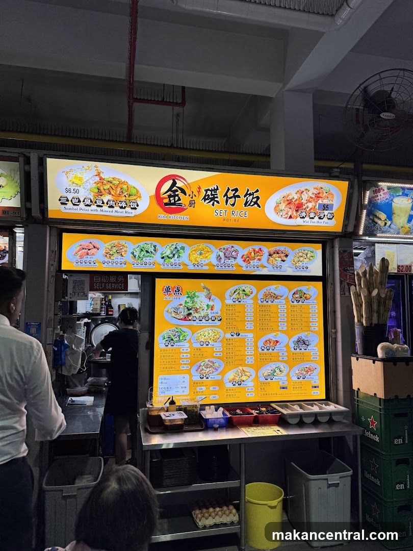 Interior view of a food stall with bright yellow signage displaying '金小厨 碟仔饭 SET RICE'. A large digital menu board shows various set rice dishes with prices. Customers are present; one is ordering, another waiting. The counter has condiments and prepared items. Stacks of Heineken crates and a fan are visible in the background.