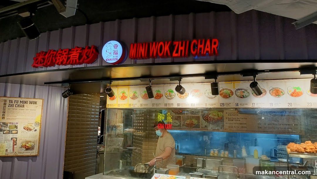Storefront with red LED signage reading '迷你锅煮炒' and 'MINI WOK ZHI CHAR', featuring a circular logo with Chinese characters. A glass-enclosed cooking area is visible behind the counter, with a staff member wearing a face mask. The wall above displays food images and numbers indicating menu items. Left side has a vertical brick panel and a framed promotional poster.