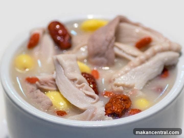 A close-up shot of a white ceramic bowl containing a light-colored broth with slices of pork offal, yellow corn kernels, and red goji berries floating in the liquid.