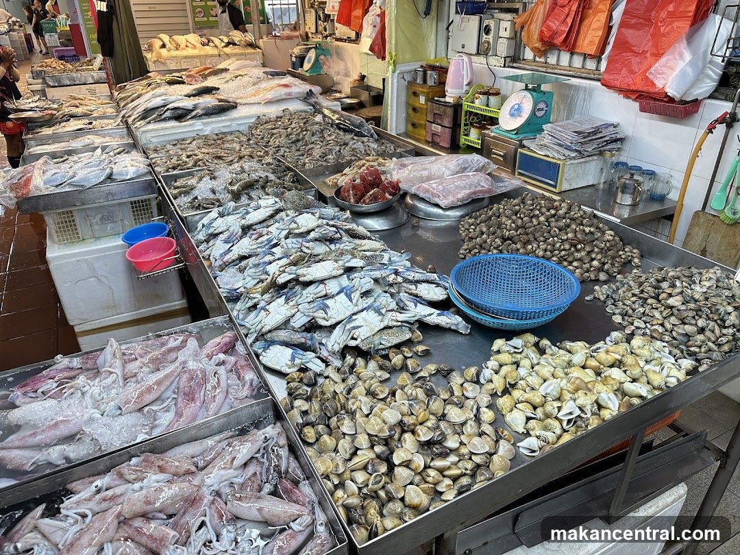 A bustling seafood market stall with various fresh fish, shrimp, crabs, clams, and squid displayed on metal trays. The counter includes weighing scales, plastic baskets, and stacked newspapers. Background shows other vendors and customers in motion.