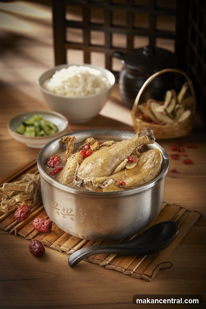 A steaming pot of herbal chicken soup garnished with red dates and ginseng sits on a bamboo mat beside rice, cucumber slices, and a basket of dried herbs. A black spoon rests nearby on wooden surface with warm lighting and traditional lattice background.