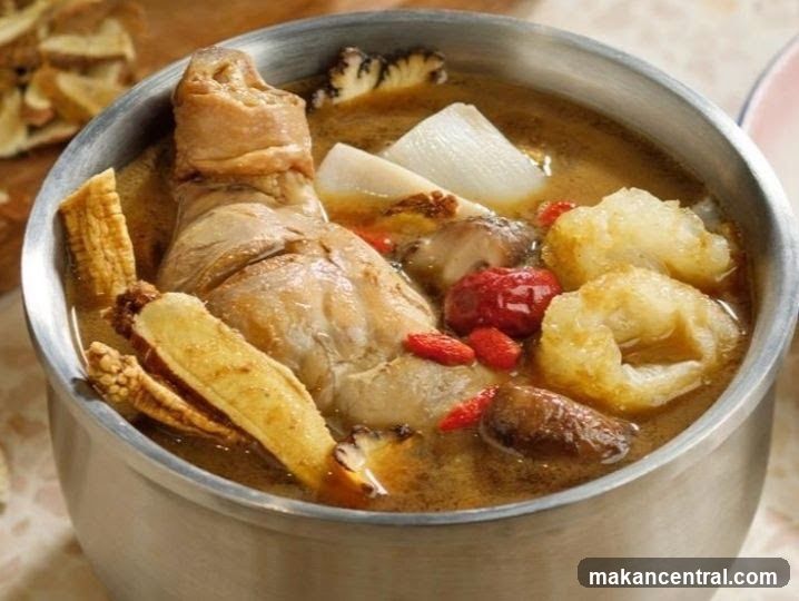 A metal bowl filled with a rich, brown broth containing chicken leg, mushrooms, goji berries, and other ingredients. The bowl sits on a light-colored surface with some dried herbs or slices nearby.