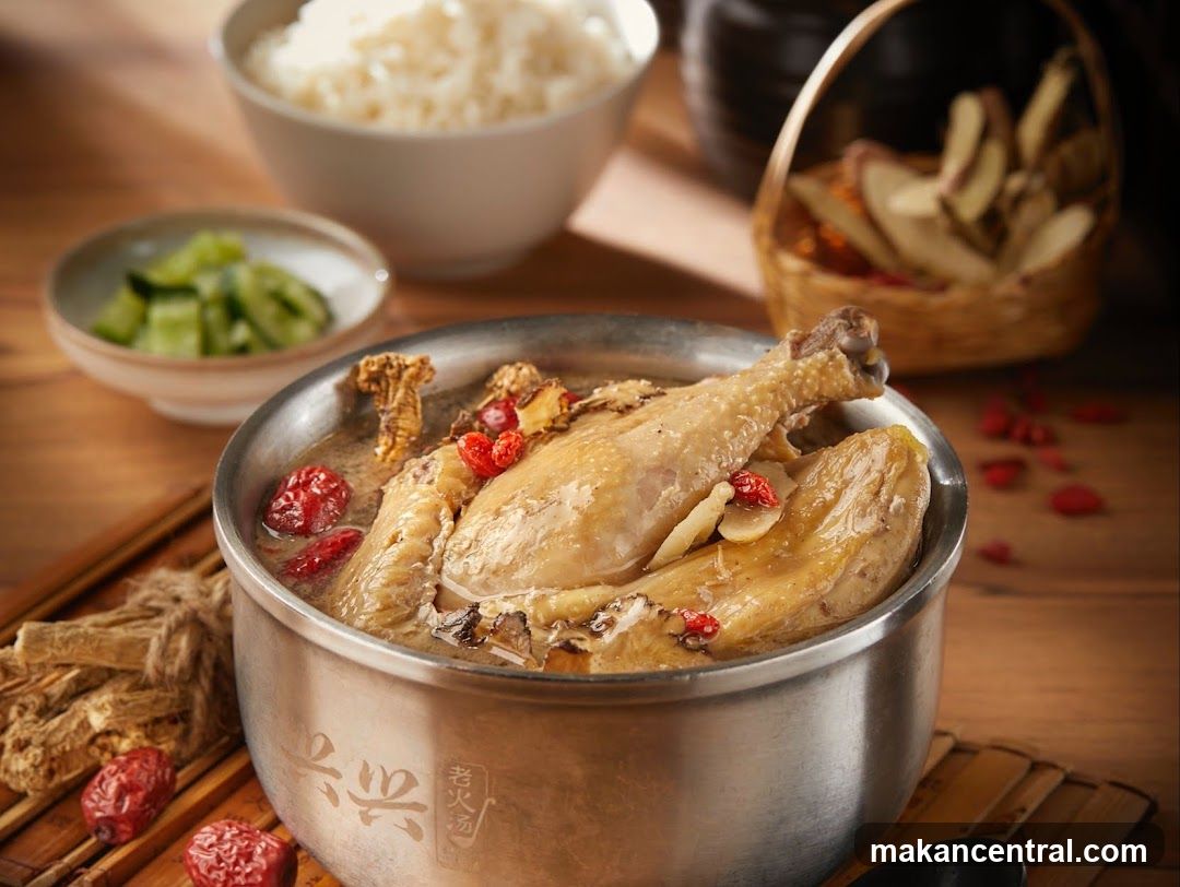 A close-up shot of a metal pot containing chicken soup garnished with goji berries and herbs, served with white rice and cucumber slices in the background. A basket of dried ingredients is also visible on a wooden table.