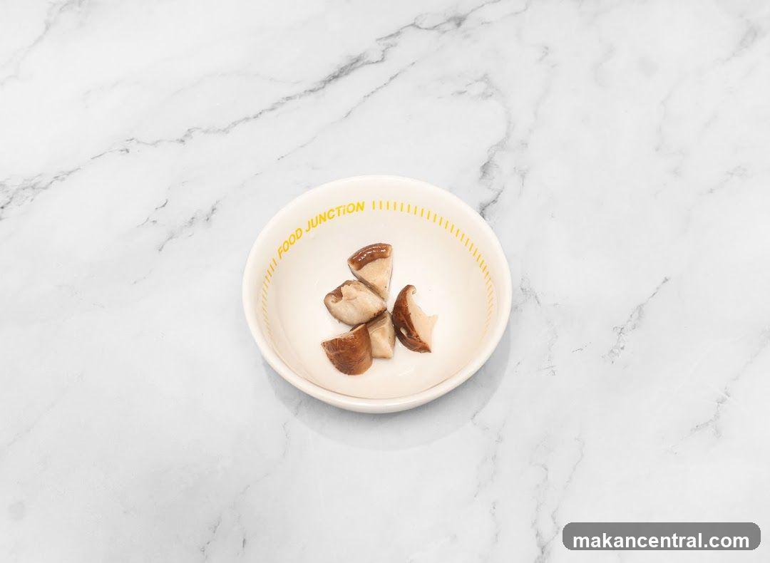 A small white ceramic bowl with yellow "FOOD JUNCTION" lettering contains several sliced mushrooms. The bowl rests on a white marble surface with gray veining.