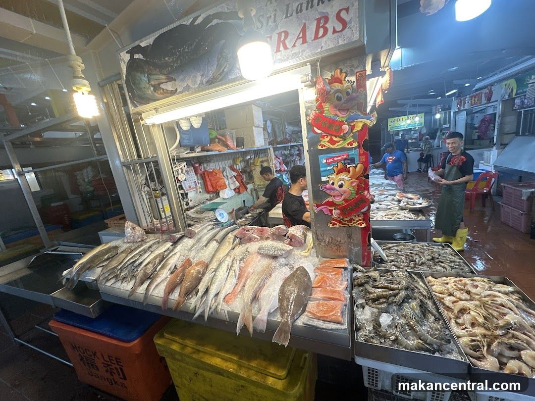 Interior view of a seafood market stall with fresh fish, shrimp, and crabs displayed on ice. Staff are present, one wearing an apron and yellow boots. A sign above reads 'Sri Lanka CRABS' with a large crab illustration. Decorative Chinese dragon motifs adorn the stall. Bright overhead lighting illuminates the display. The floor is wet, suggesting recent activity or rain. Various containers and equipment are visible in the background.