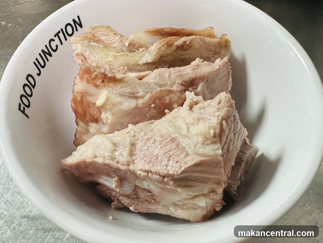 A white bowl with "FOOD JUNCTION" printed on the side contains several pieces of cooked meat, likely pork or beef, with visible fat and bone. The bowl sits on a metallic surface.