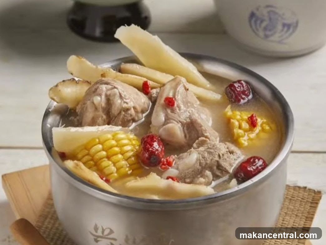 A metal pot filled with a clear broth containing pork bones, corn kernels, red dates, and sliced bamboo shoots. The pot rests on a woven mat with a wooden spoon beside it. In the background, there is a blurred ceramic bowl with a blue design and part of a dark object. The setting suggests an indoor dining or kitchen environment.