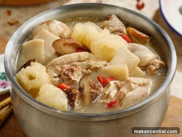 A close-up shot of a metal bowl containing a light-colored broth with chunks of meat, mushrooms, and red goji berries. The bowl sits on a wooden surface with partial view of a plate and other kitchenware in the background.