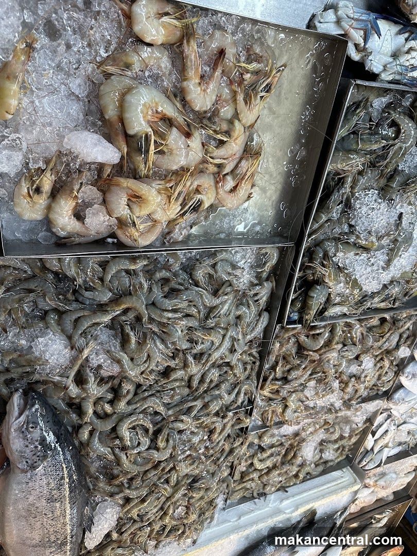 Fresh seafood including shrimp, fish, and crab displayed on ice in metal trays. Shrimp are arranged in various sizes and states of freshness, with some larger, peeled specimens and smaller whole ones. Ice is visibly melting around the products. The display appears to be part of a wet market or seafood stall.