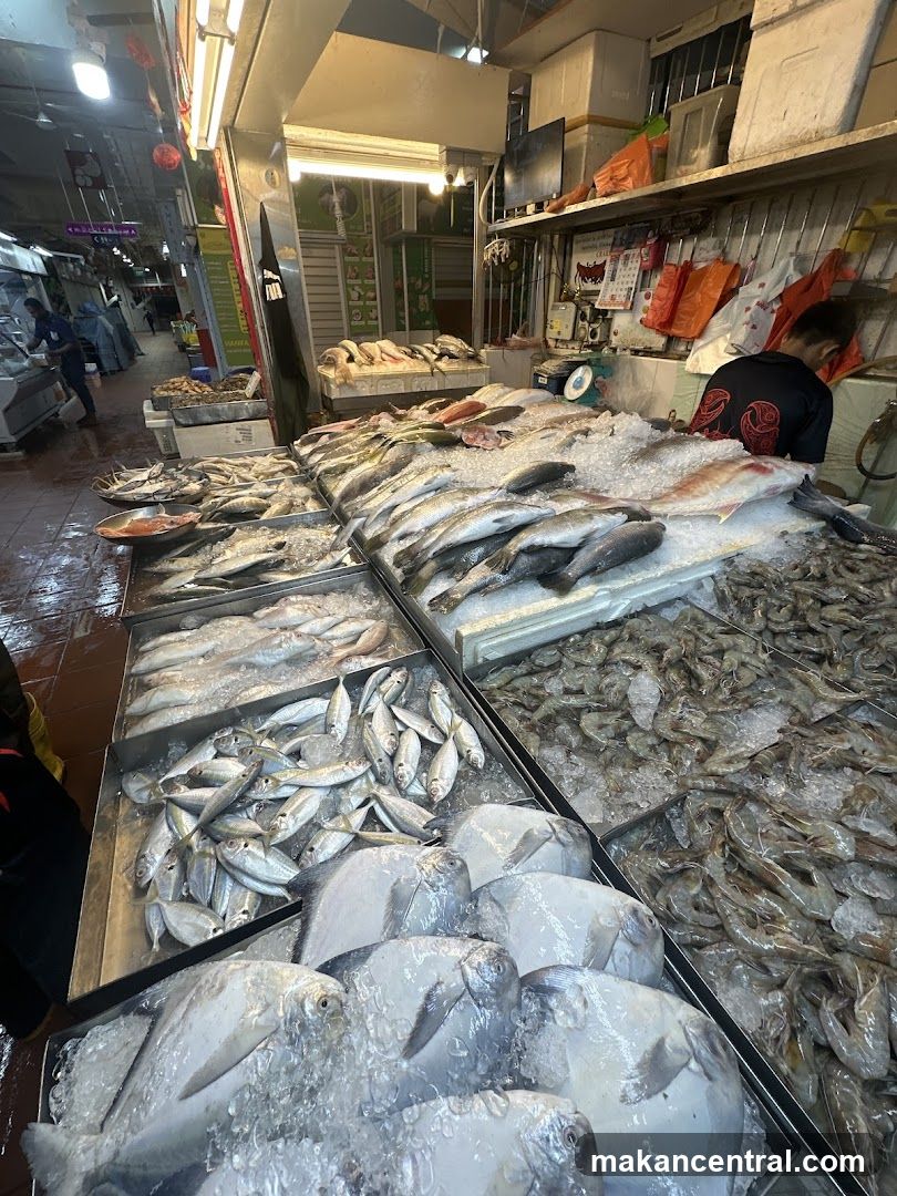 Interior view of a fish market stall with various seafood displayed on ice. Multiple types of fish and shrimp are arranged in trays. A vendor is present behind the counter. The floor appears wet, and overhead lighting illuminates the area. Background shows other stalls and shoppers.