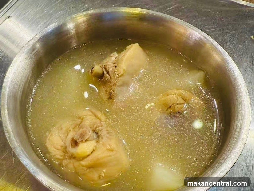A metal bowl containing a light-colored broth with several pieces of boiled meat submerged in it. The bowl rests on a metallic surface, likely a countertop or table. No signage, storefront, or printed materials are visible.