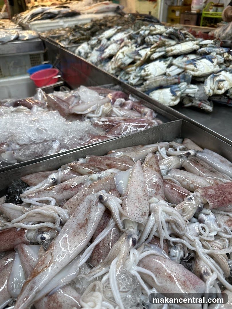 Fresh seafood displayed on ice in metal trays, including squid and crabs. Ice is visible beneath the fish. Background shows more seafood and market stalls with plastic containers. No signage or storefront visible.
