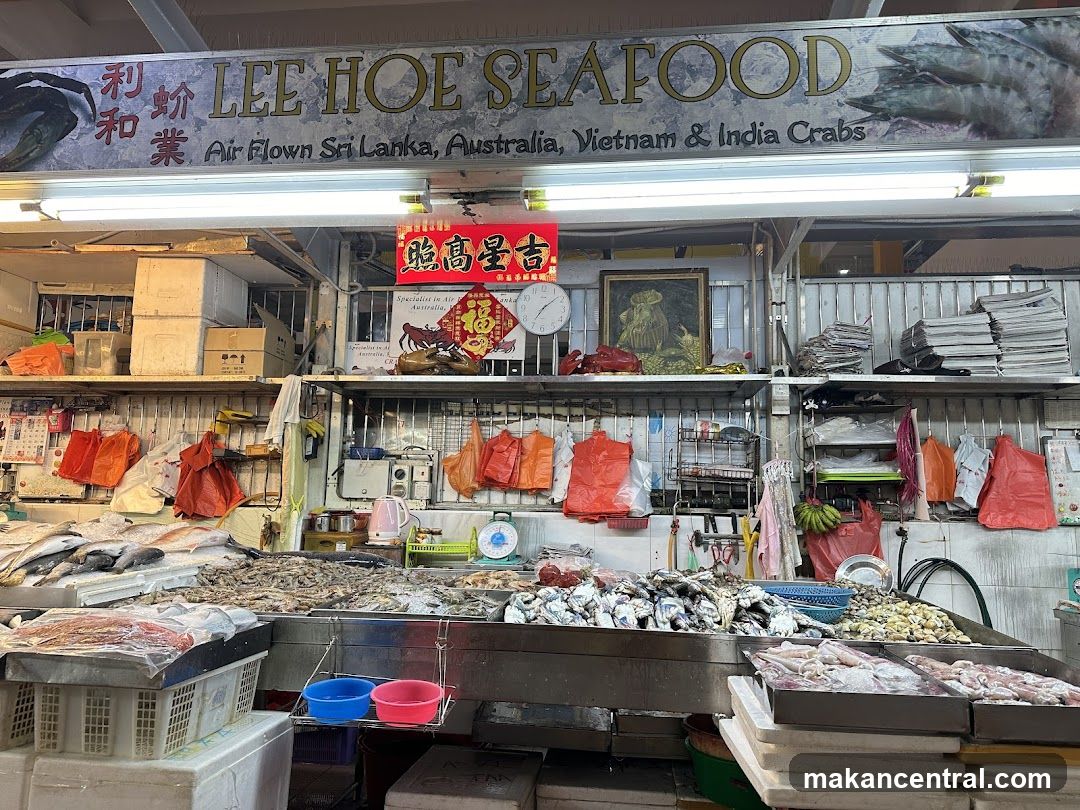 Interior view of a seafood stall with fresh fish and shellfish displayed on metal counters. Above the counter is a sign reading 'LEE HOE SEAFOOD' with Chinese characters to the left and text below listing 'Air Flown Sri Lanka, Australia, Vietnam & India Crabs'. Red decorative banners hang above the display area. Shelves hold boxes, bags, and kitchen equipment. A clock and framed picture are visible on the wall behind the counter.