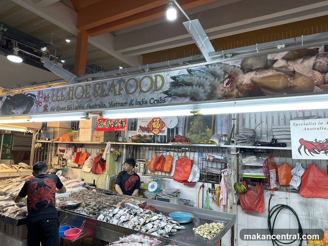 Interior view of a seafood stall with staff working behind counters displaying fresh fish and shellfish. Signage above reads 'LEE HOE SEAFOOD' and advertises crabs from Sri Lanka, Australia, Vietnam & India. Red plastic bags hang on racks, and promotional posters are visible. The ceiling has exposed beams and lighting fixtures.