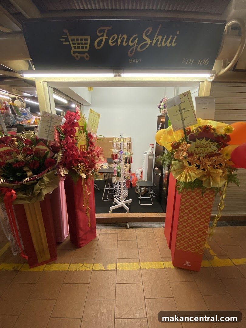 Storefront entrance with illuminated sign displaying 'Feng Shui MART' and unit number #01-106. Two large floral arrangements flank the doorway; one labeled 'BEST WISHERS', another with Chinese characters. Interior shows retail display racks, tables, and chairs under fluorescent lighting. Yellow floor marking lines are visible in foreground.