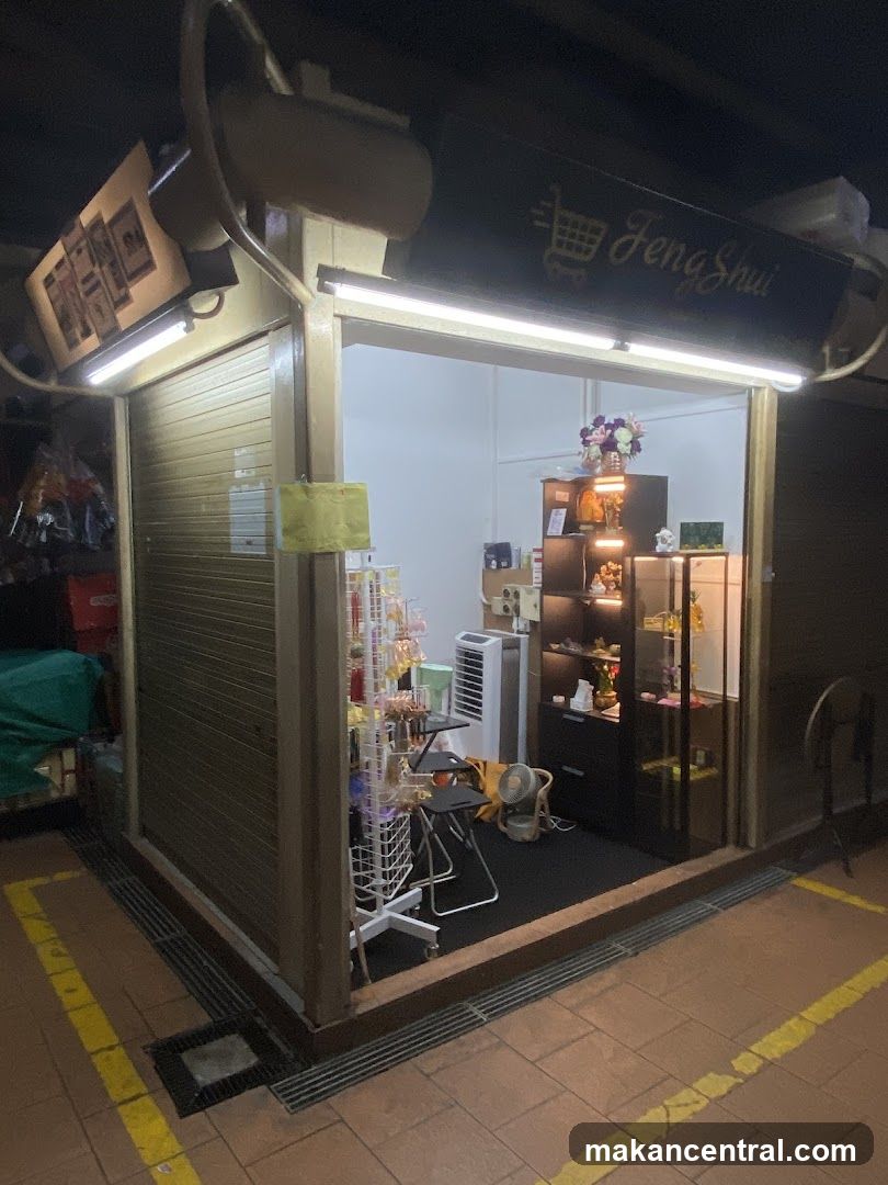 Nighttime exterior view of a storefront with illuminated signage reading 'Feng Shui' featuring a shopping cart icon. The shop has a partially open entrance revealing interior displays including shelves, a glass cabinet, an air conditioner, and decorative items like flowers. A yellow sticky note is affixed to the door frame. The ground floor has tiled flooring with yellow painted lines and a drainage grate.