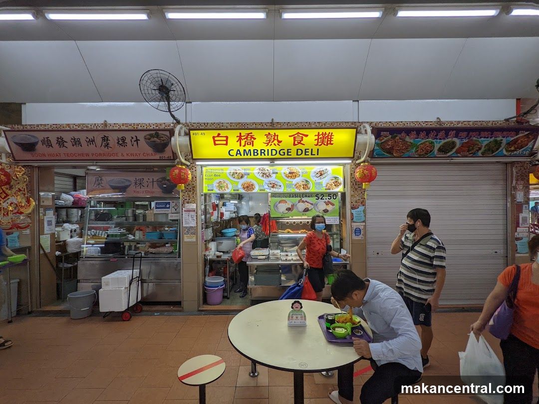 Interior view of a food stall with customers and staff wearing masks. A man eats at a table in the foreground while others stand nearby. The stall has bright yellow signage reading '白橋熟食攤 CAMBRIDGE DELI'. Adjacent stalls are visible, one labeled '順發潮州糜煩汁' (Soon Hua Teochew Porridge & Kway Chap). Red lanterns hang above. A fan is mounted on the ceiling.
