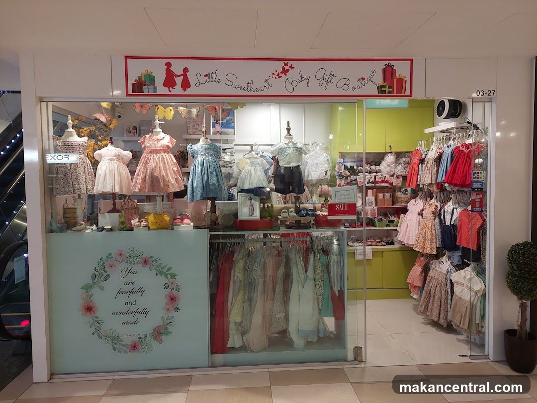 Storefront with glass display windows showcasing baby clothing on mannequins and racks. Above the entrance is a red-bordered sign with the shop name and decorative elements like gift boxes and silhouettes. A frosted panel on the left features floral wreath design with text: 'You are fearfully and wonderfully made.' A small 'SALE' sign is visible inside. The store interior has bright lighting, lime green cabinets, and hanging garments. To the right, a potted plant stands beside the entrance.