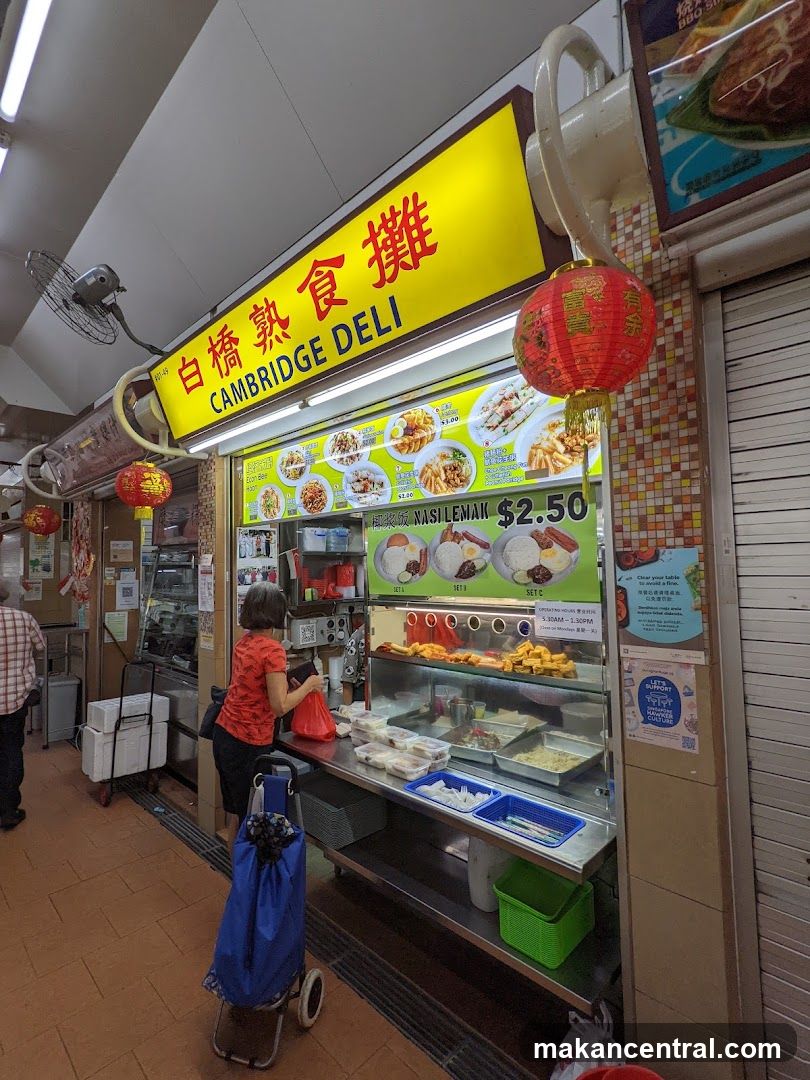 Interior view of a food stall with bright yellow signage reading '白橋熟食攤 CAMBRIDGE DELI'. A customer in a red shirt stands at the counter with a shopping bag, while a display case holds prepared foods and a menu board shows prices like $2.50 for Nasi Lemak. Red lanterns hang beside the stall. The floor is tiled, and a fan is mounted on the ceiling.
