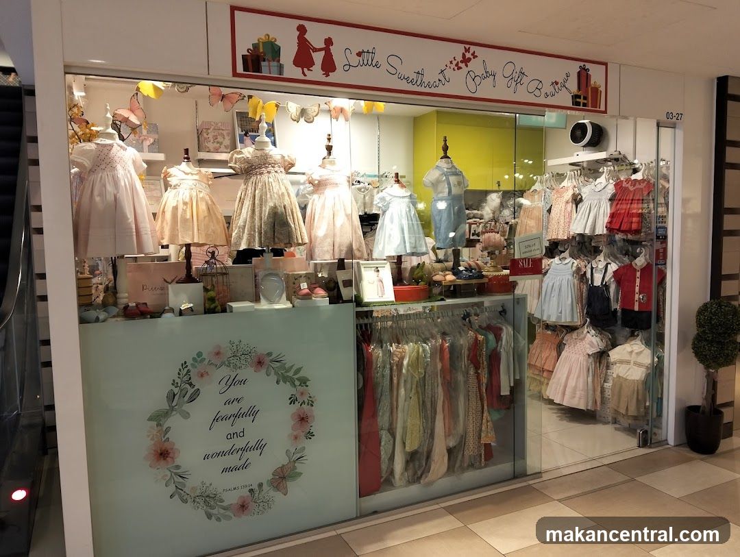 Storefront with glass display windows showcasing baby dresses on mannequins and racks of clothing. Signage above reads 'Little Sweetheart Baby Gift Boutique' with decorative elements like butterflies and gift boxes. A frosted panel below the window features a floral wreath design with the text 'You are fearfully and wonderfully made'. The shop number 03-27 is visible on the right side. Interior lighting illuminates the merchandise, and a small potted plant stands beside the entrance.