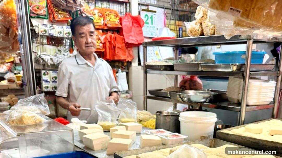 An elderly man stands behind a counter in a bustling tofu shop, surrounded by stacks of white tofu blocks, metal trays, and shelves stocked with packaged goods. The background includes red shopping bags, plastic containers, and hanging food products. A calendar with the number '21' is visible on the wall.