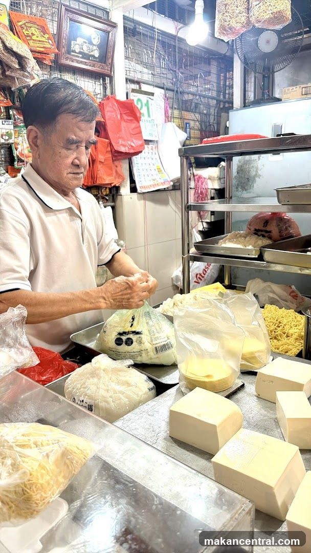 An elderly man in a white polo shirt is preparing food behind a counter filled with packaged noodles, tofu blocks, and other ingredients. The setting appears to be a small food stall or kitchen with metal shelving, hanging fans, and various printed materials on the walls including calendars and framed pictures. No shop front signage is visible from this interior perspective.