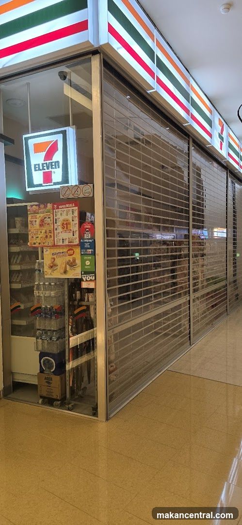 Storefront with metal roll-down shutters partially open, revealing a glass entrance. The 7-Eleven logo is prominently displayed on the facade above the entrance. Inside, shelves are stocked with products, including bottled water and snacks. A small display cart holds promotional materials. The floor is tiled in beige, and the ceiling has recessed lighting.
