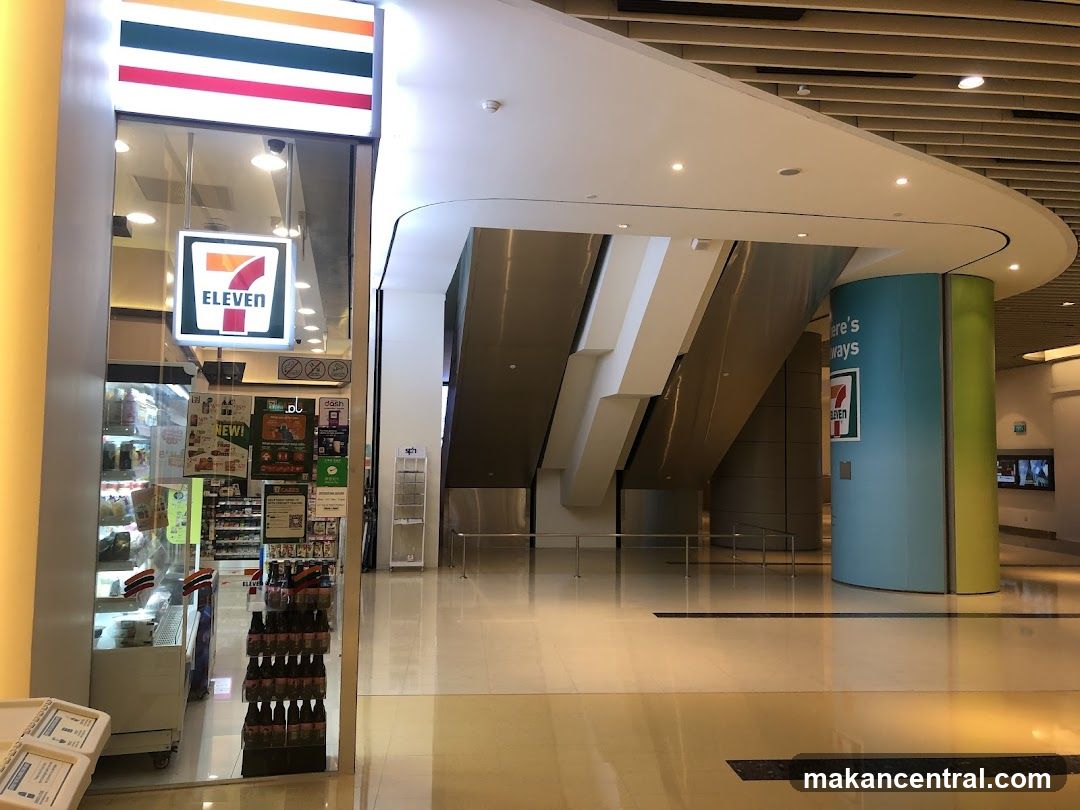 A 7-Eleven store entrance with illuminated signage above the glass facade showing the iconic red, white, and blue logo. The interior is partially visible through the transparent doors, revealing shelves stocked with products and promotional displays inside.