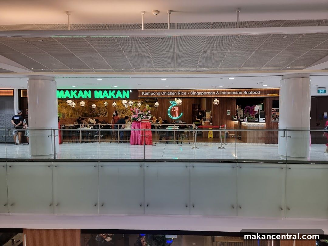 The exterior of a restaurant with a prominent green sign that reads "MAKAN MAKAN" and smaller text indicating "Kampong Chicken Rice • Singaporean & Indonesian Seafood". The restaurant has a wooden interior with tables and chairs visible, and people dining. There is a counter area with food displays and a menu board.