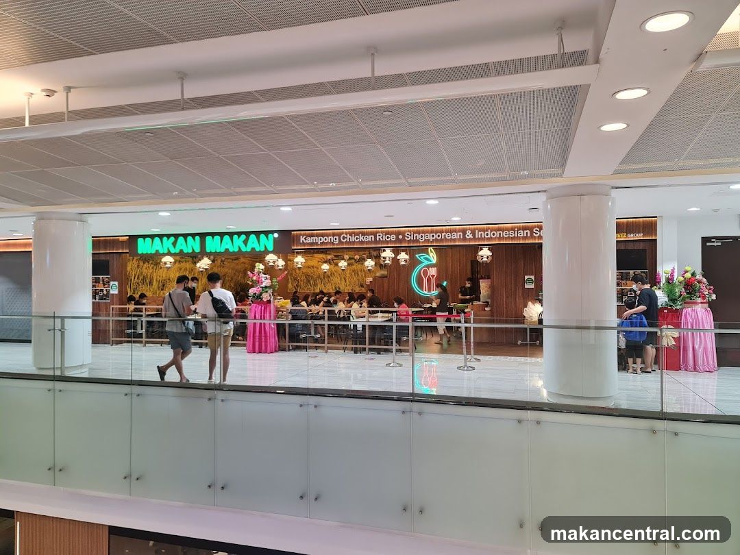 The exterior of a restaurant called Makan Makan, which serves Kampong Chicken Rice, Singaporean & Indonesian food. The restaurant has a green illuminated sign with the name "MAKAN MAKAN" and a logo of a stylized leaf with a fork and spoon. The restaurant is busy with customers seated at tables inside. There are floral decorations outside the entrance.