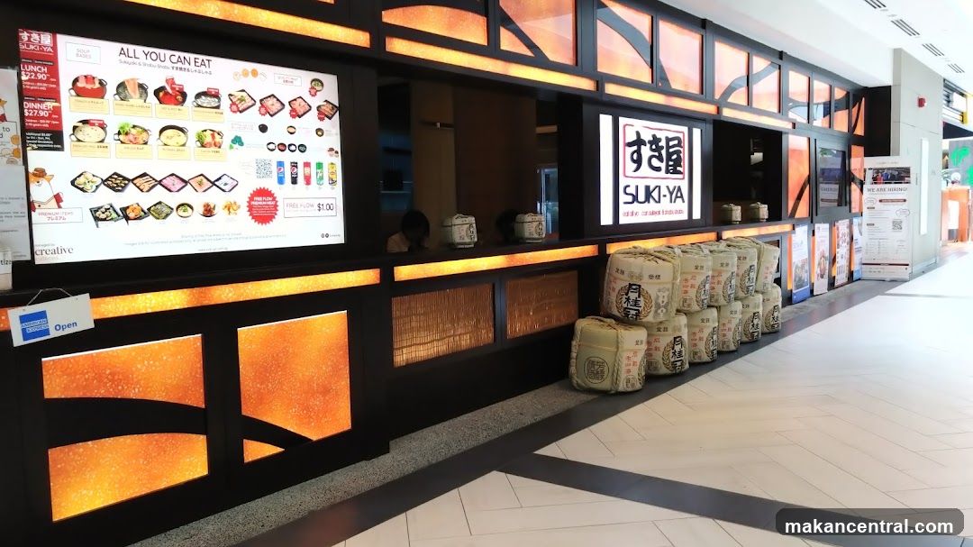 The exterior of a Suki-Ya restaurant with a large illuminated menu board displaying "All You Can Eat" options. The menu shows various soup bases, premium items, and drinks. The restaurant's logo, "SUKI-YA," is visible on the facade. Stacks of decorative sake barrels are placed outside the establishment. An "Open" sign is displayed on the counter.